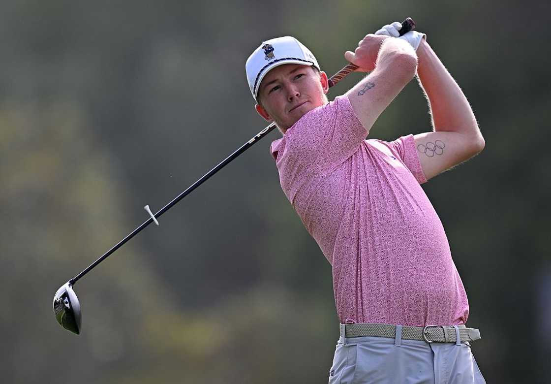 A man in a pink patterned polo and white cap watches his golf shot, driver raised behind him.
