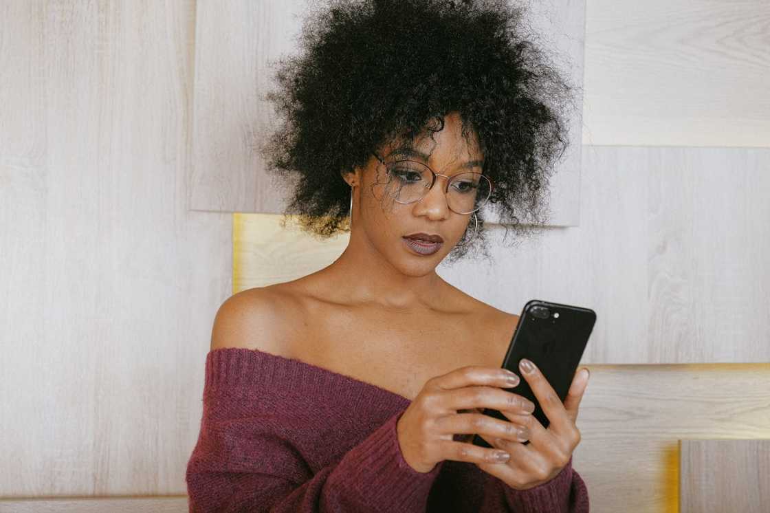 A woman wearing glasses looks down at her phone while standing against a light wooden wall. A woman wearing glasses looks down at her phone while standing against a light wooden wall.