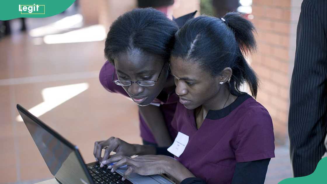 A woman in glasses leans over another woman's shoulder as she types on a laptop at a wooden table.