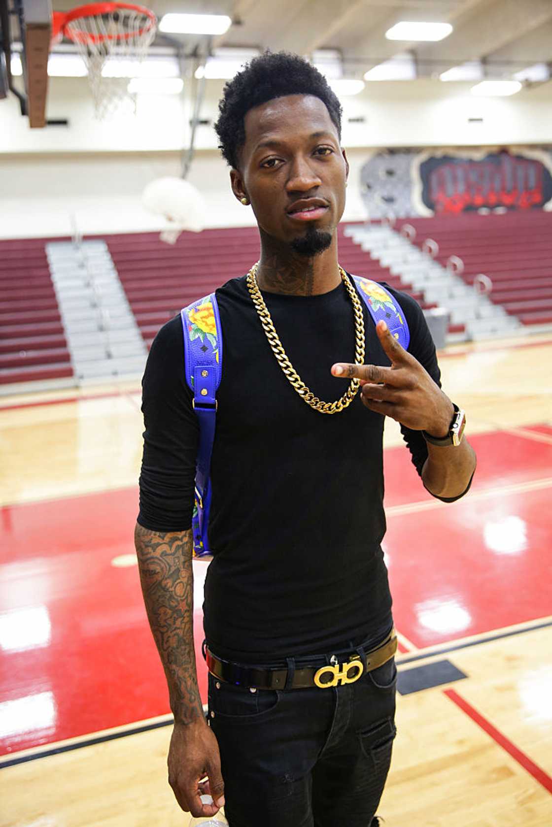 A man in a black long-sleeve shirt and gold chain poses with a peace sign on an indoor basketball court. A man in a black long-sleeve shirt and gold chain poses with a peace sign on an indoor basketball court.
