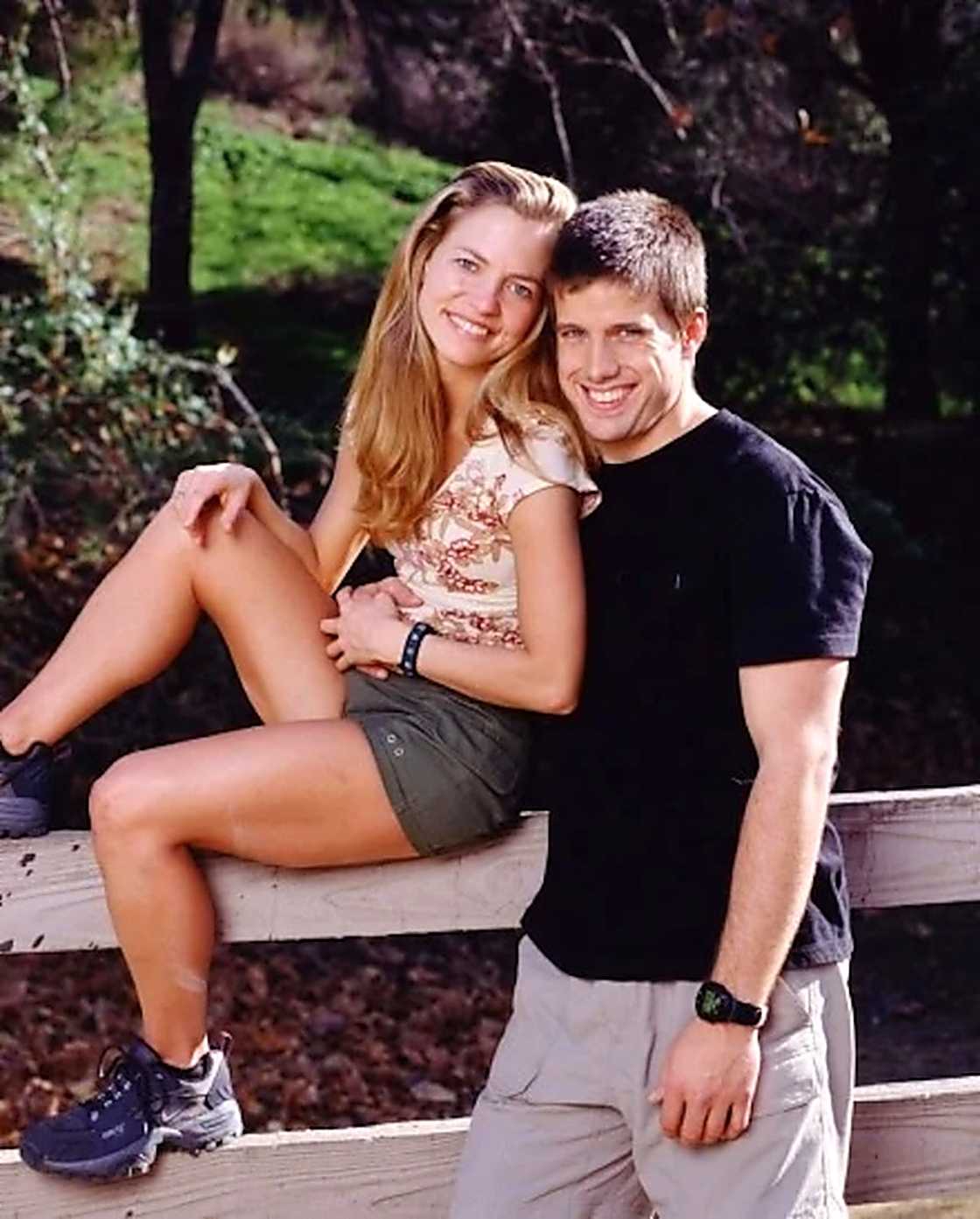 A woman with long blonde hair sits on a wooden fence rail, leaning against a man in a black t-shirt.