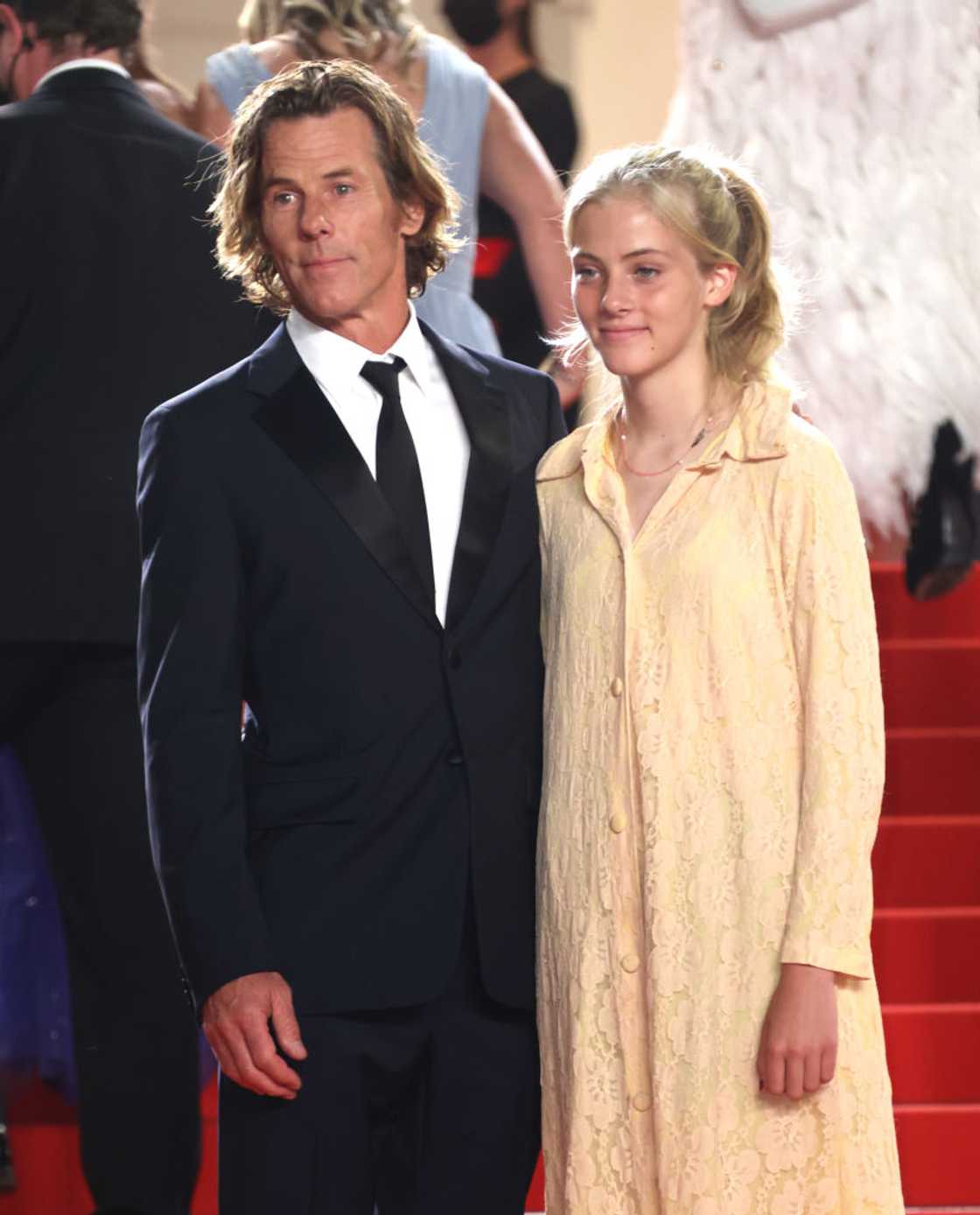 A man in a black tuxedo stands beside a teenage girl in a pale yellow lace dress on a red carpet. A man in a black tuxedo stands beside a teenage girl in a pale yellow lace dress on a red carpet.