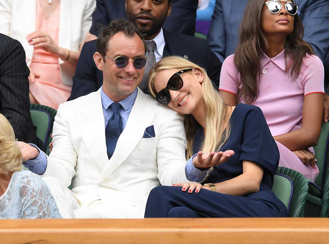 Jude Law in a white suit sits with a woman resting her head on his shoulder in the Wimbledon stands.