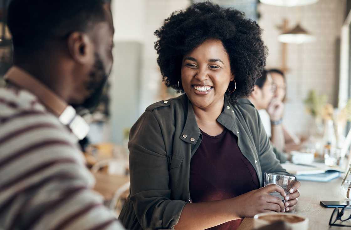 Two people sit at a café counter, one smiling and holding a glass of water.