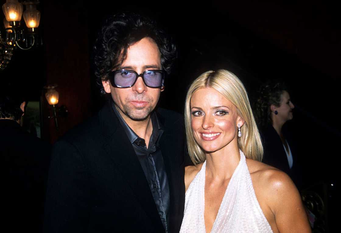 A man in sunglasses and a dark blazer stands with a woman in a white halter dress indoors. A man in sunglasses and a dark blazer stands with a woman in a white halter dress indoors.