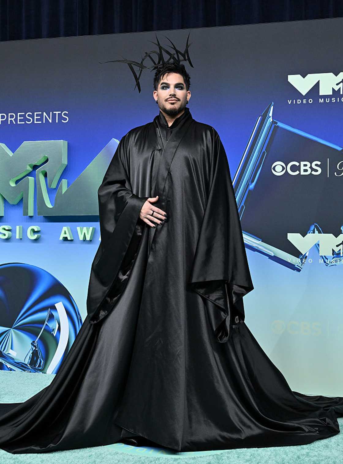 Adam Lambert with dark swept hair and bold eye make-up in a black satin cape, smiling at the camera at the 2026 MTV Video Music Awards at Peacock Theater in Los Angeles