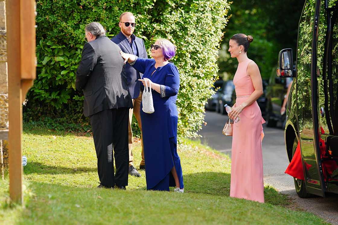 Three men and two women in formal wear stand on grass near a car outside a hedge-lined venue.
