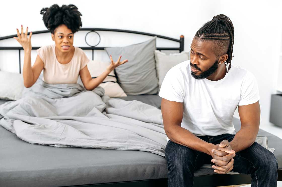 A woman scolds her upset husband sitting on the bed