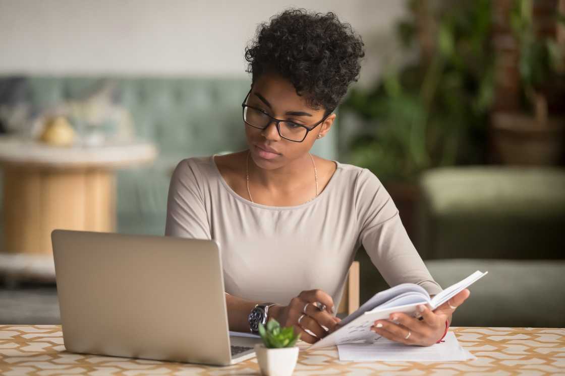 Focused young businesswoman looking at laptop holding book learning