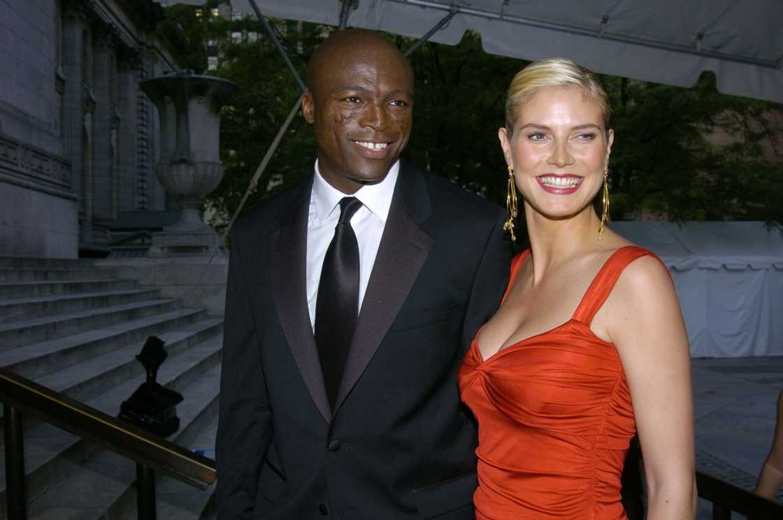 A man in a black tuxedo with a white shirt and black tie stands next to a woman with slicked-back blonde hair wearing a bright orange satin gown. A man in a black tuxedo with a white shirt and black tie stands next to a woman with slicked-back blonde hair wearing a bright orange satin gown.