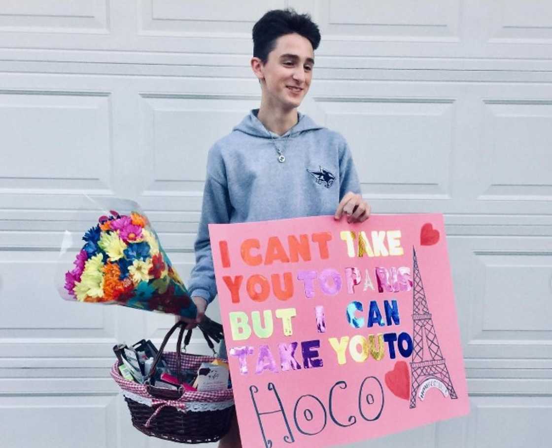 A young man holding a Homecoming poster, flowers and a gift hamper