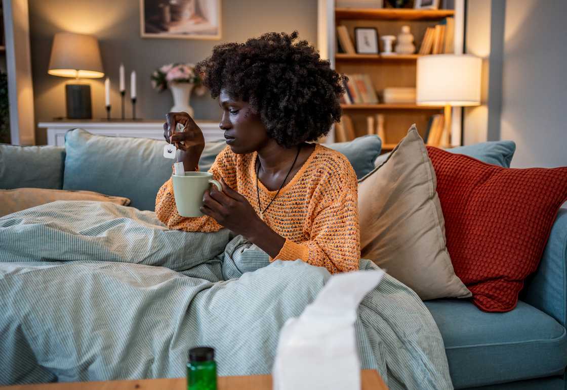 Person on couch with blanket and tea, medicine and tissues nearby. Person on couch with blanket and tea, medicine and tissues nearby.