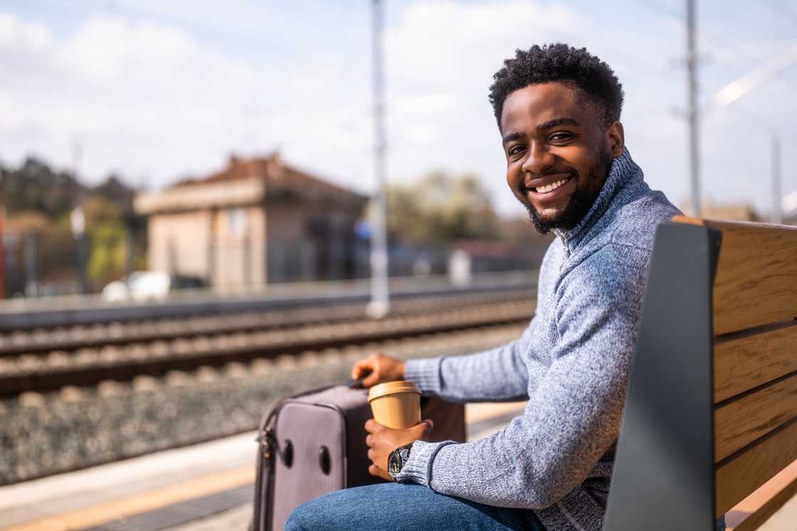 Person sits on a wooden bench at a train station, smiling and holding a coffee cup with a suitcase beside them.