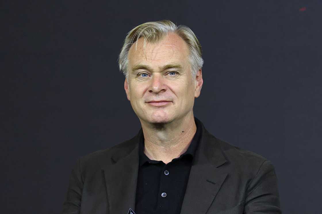 Christopher Nolan wearing a black blazer over a black polo shirt, smiling slightly against a dark background. Christopher Nolan wearing a black blazer over a black polo shirt, smiling slightly against a dark background.
