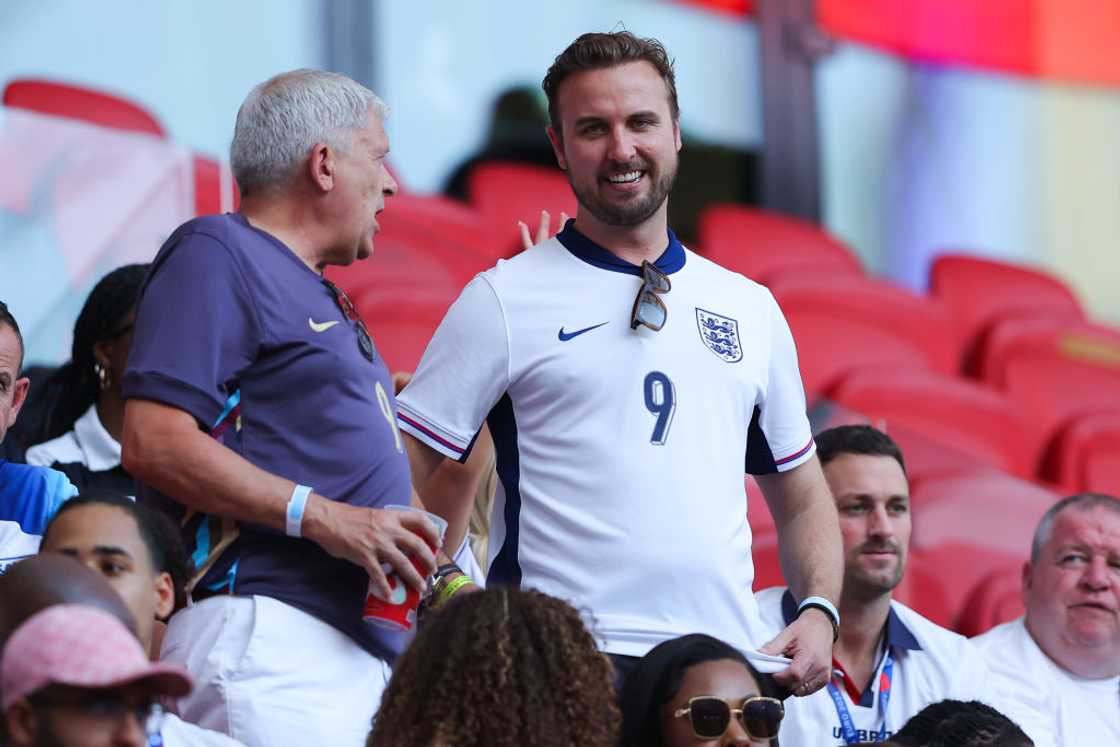 A man in a white England jersey smiles while talking to an older man in a purple shirt. A man in a white England jersey smiles while talking to an older man in a purple shirt.