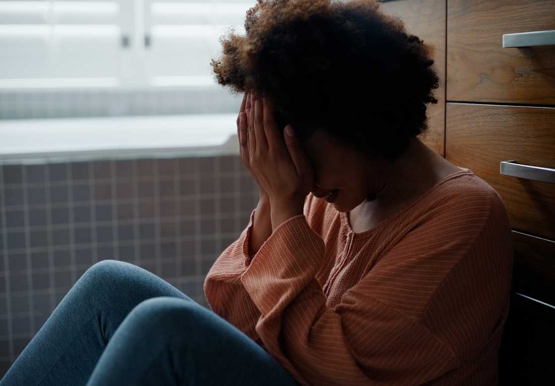 Person sits on the floor with head in hands, leaning against a cabinet in a softly lit bathroom. Person sits on the floor with head in hands, leaning against a cabinet in a softly lit bathroom.