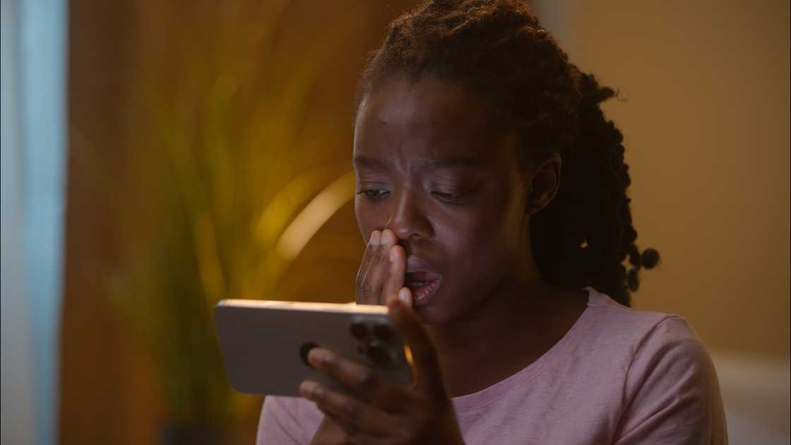 A person reacts with hand over mouth while looking at a smartphone indoors.