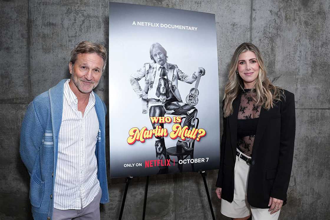 A man in a blue cardigan and a woman in a black blazer stand beside a film poster on an easel. A man in a blue cardigan and a woman in a black blazer stand beside a film poster on an easel.