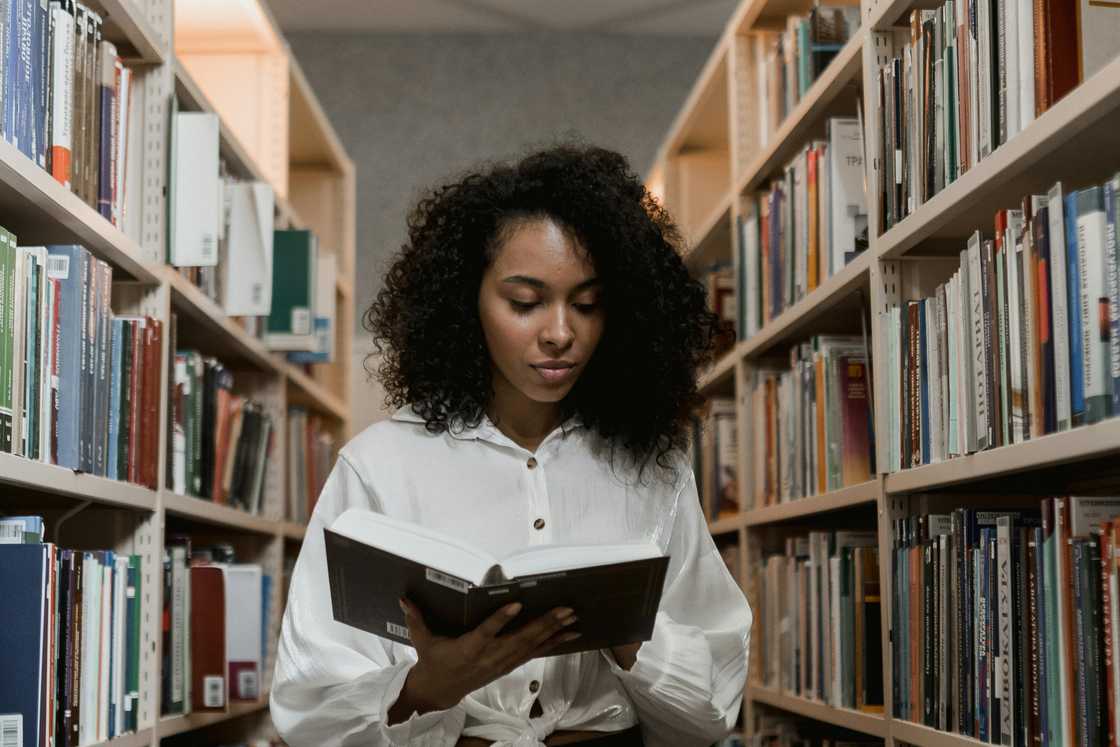 A young woman in a white button shirt is reading a book in the library