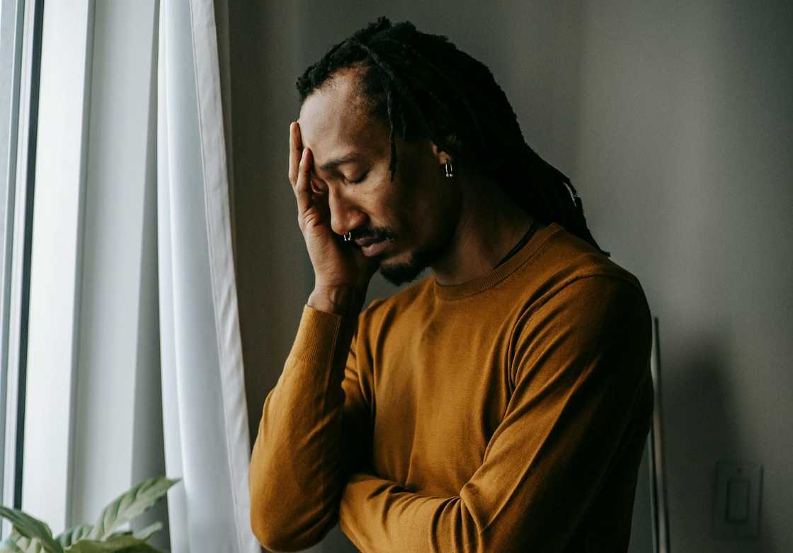 A grieving man stands by a window with his hand on his forehead, eyes closed in quiet distress. A grieving man stands by a window with his hand on his forehead, eyes closed in quiet distress.