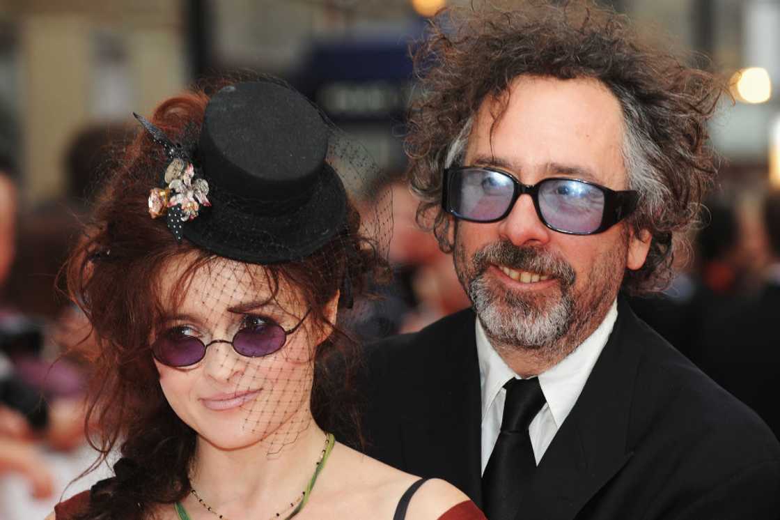 A curly-haired man in glasses stands close to a woman in a veiled black hat and dark top.