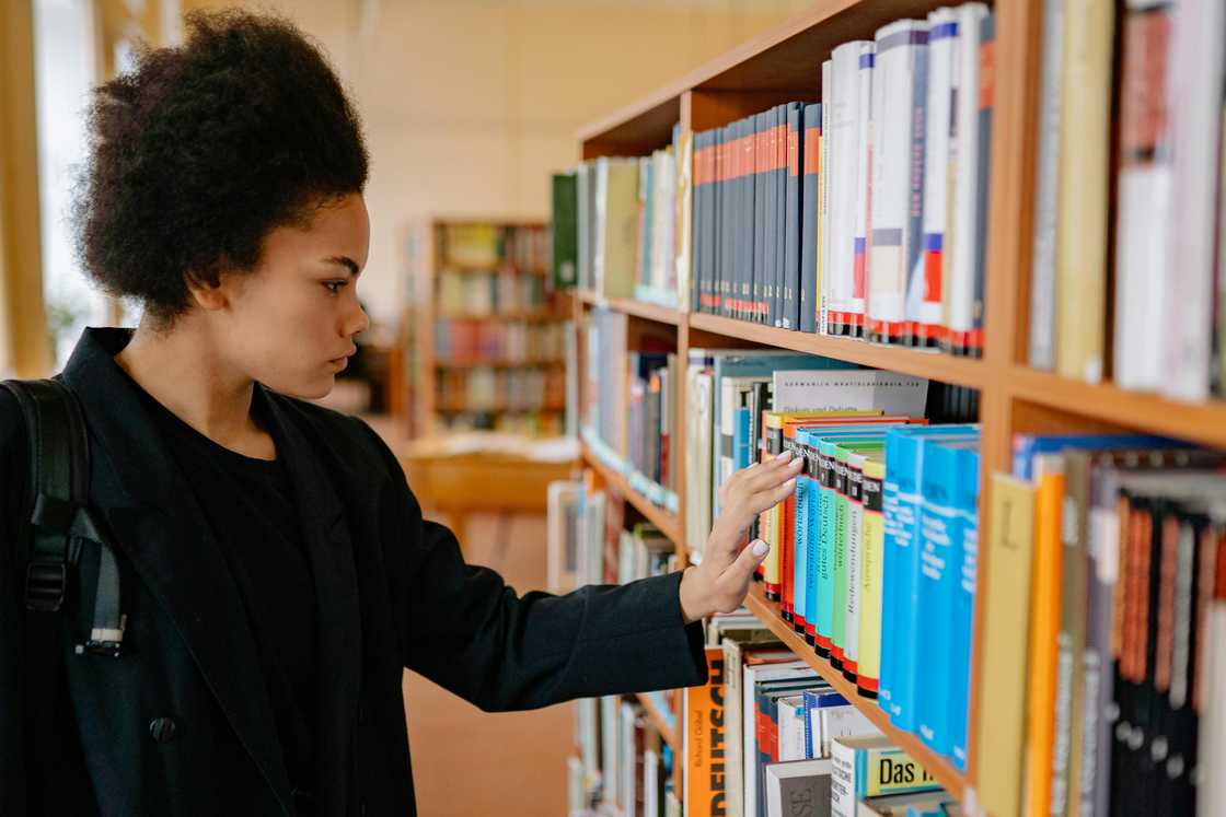 A young woman in Afro hair and a black outfit is searching for a book in a library