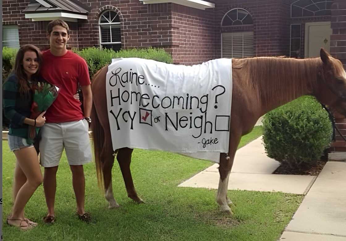 A couple posing for a photo next to a house draped with a Homecoming proposal banner