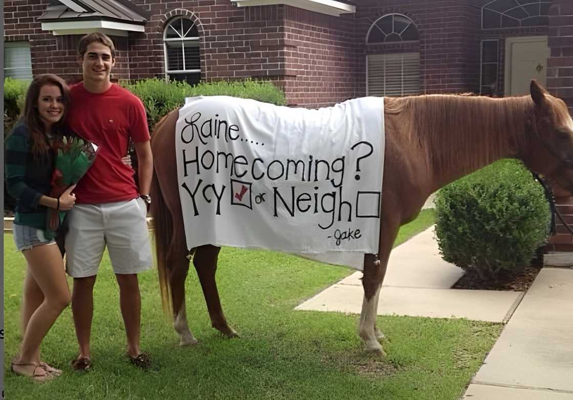 A couple posing for a photo next to a house draped with a Homecoming proposal banner A couple posing for a photo next to a house draped with a Homecoming proposal banner