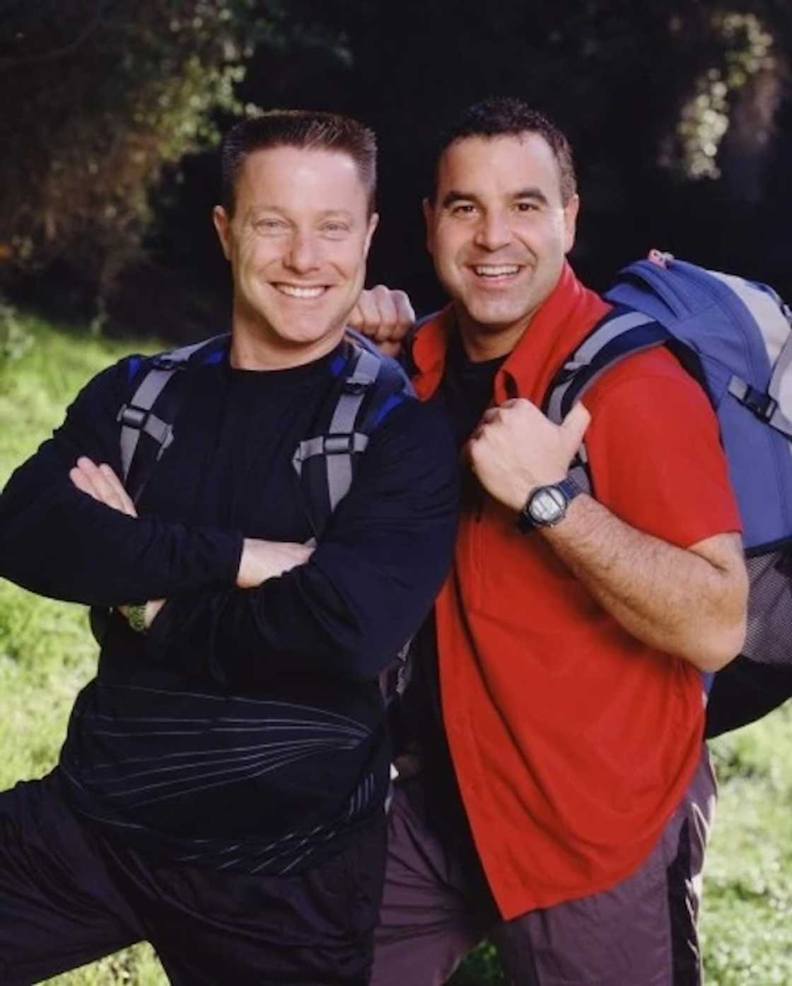A man in a black long-sleeve top crosses his arms and poses next to a man in a red vest giving a thumbs up. A man in a black long-sleeve top crosses his arms and poses next to a man in a red vest giving a thumbs up.