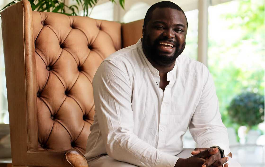 Iyinoluwa Aboyeji smiles broadly while seated in a brown button-tufted armchair.
