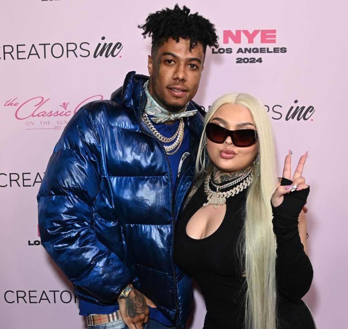 Blue Face and Jaidyn Alexis pose in a black cutout top, sunglasses, and long platinum blonde hair at a pink "NYE Los Angeles 2024" step-and-repeat event.