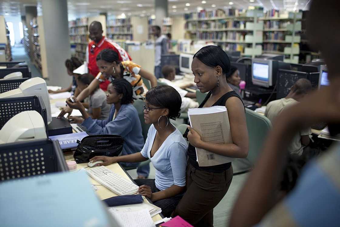 Several students sit and stand at rows of desktop computers in a library, with one woman holding papers and folders.