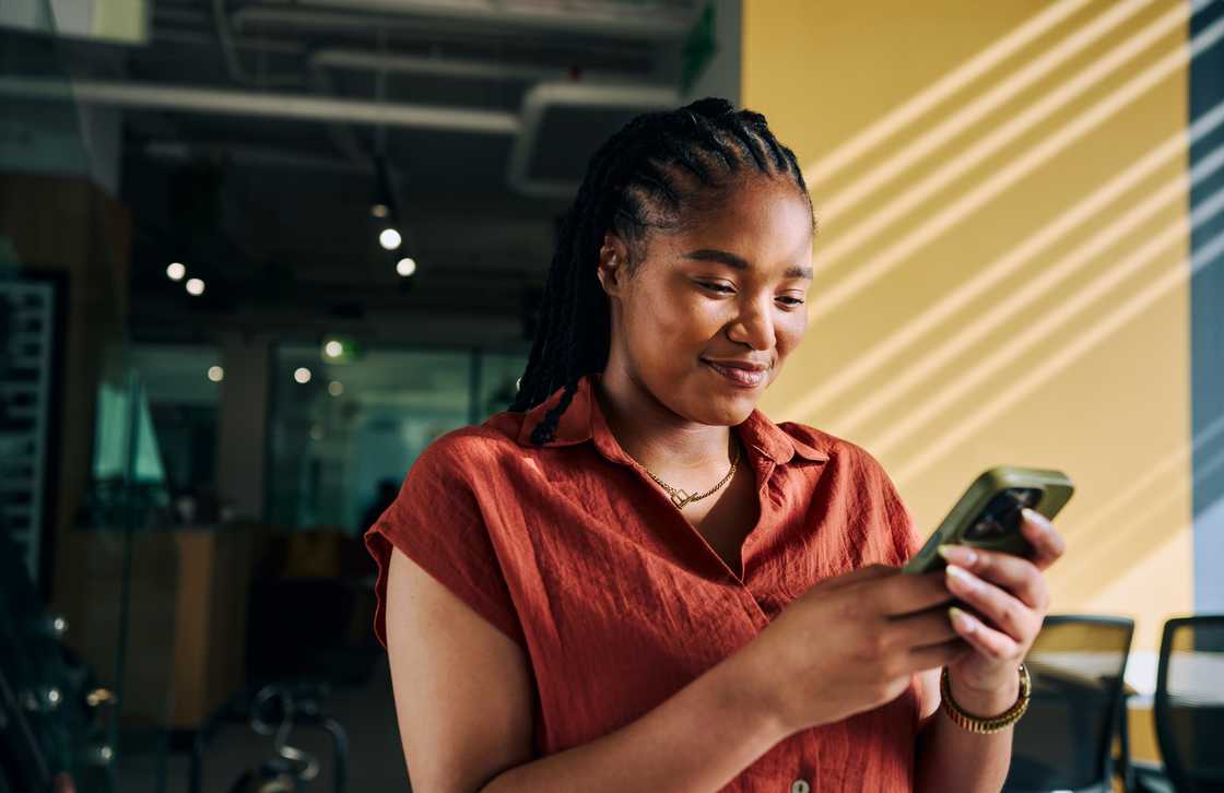 A woman with braided hair and a red blouse looks down at her phone.