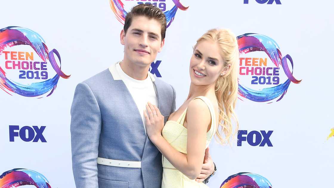 Gregg Sulkin, in a grey coat and Michelle Randolph, in a yellow dress, hold each other on the red carpet of the 2019 Fox Teen Choice Awards