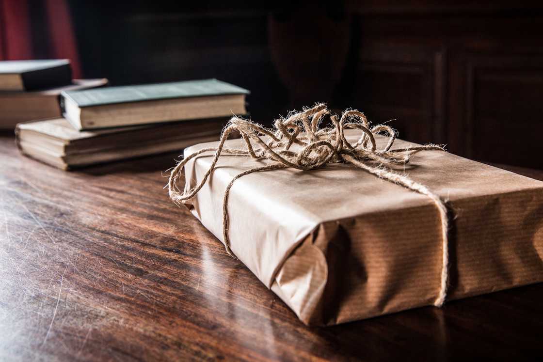 Brown paper-wrapped package tied with twine sits on a wooden table beside stacked hardcover books.