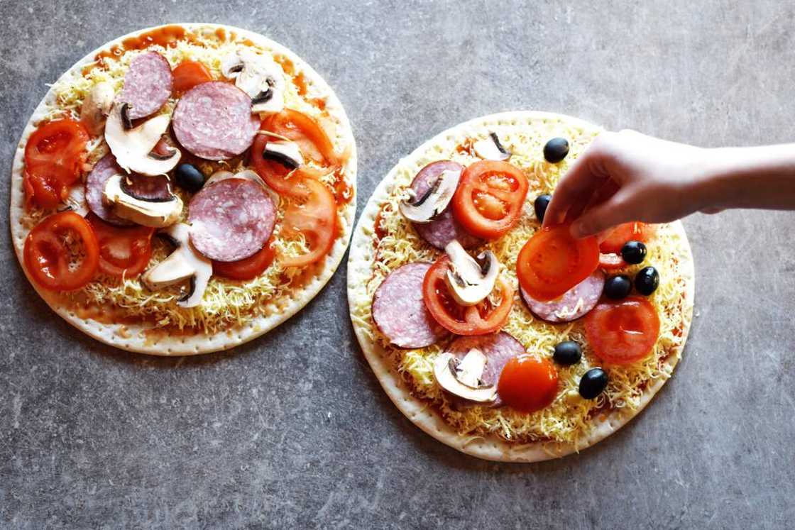 Teenage girl's hand putting slice of tomato on raw pizza with tomato, cheese, salami, olives and mushrooms ready for baking.