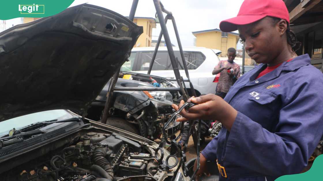 Mechanical engineer Ajaye Oluwatosin in navy overalls and a red cap, working on a car engine and facing the camera at her workshop in Lagos, 2023