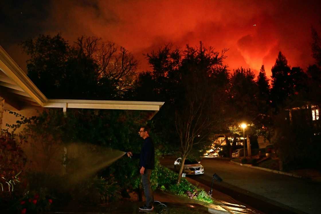 A man waters the front of his house as smoke and flames from the Palisades Fire burn toward the Encino neighborhood of Los Angeles