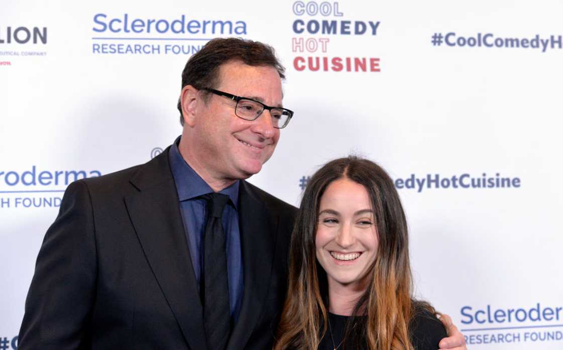A man in a black suit and glasses smiles beside a young woman with long dark hair. A man in a black suit and glasses smiles beside a young woman with long dark hair.
