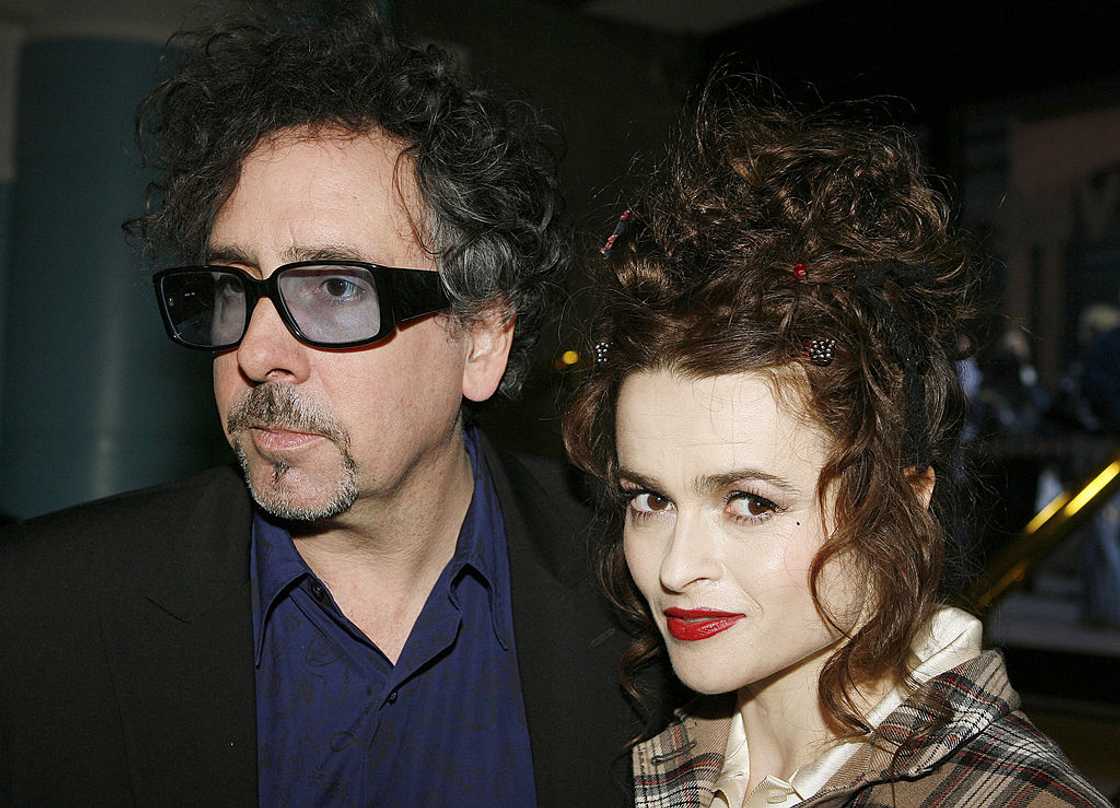 A man in a dark coat and sunglasses stands beside a woman with curly hair in a plaid coat.