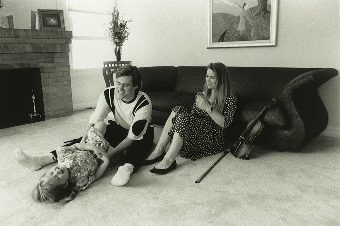 A black-and-white photo of Jim Carrey sitting on the floor with a young girl, a woman seated on a couch nearby with a violin beside her. A black-and-white photo of Jim Carrey sitting on the floor with a young girl, a woman seated on a couch nearby with a violin beside her.