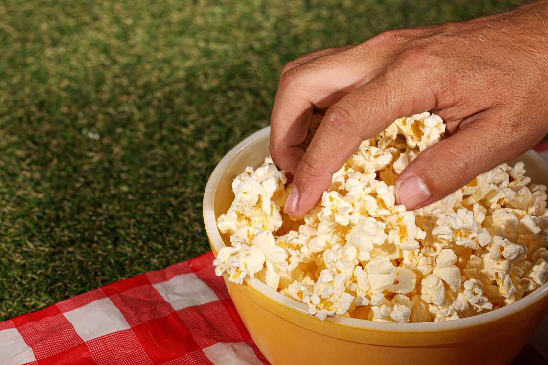 A close-up shot of a hand reaching into a bowl of popcorn on a picnic blanket on a patch of grass