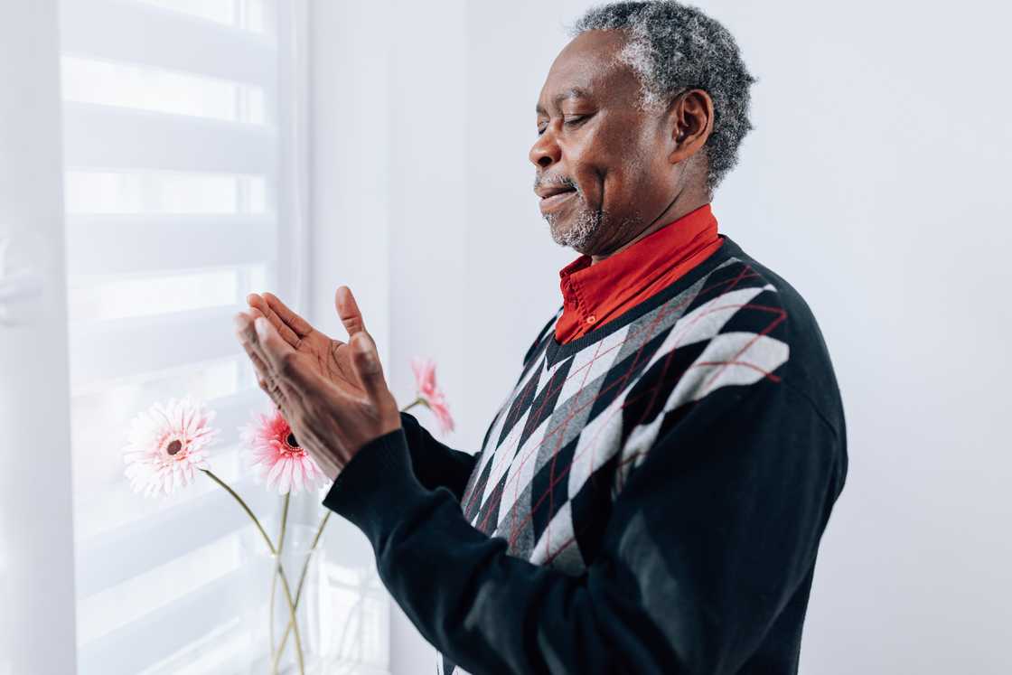 Older person stands indoors near a window with hands gently clasped. Older person stands indoors near a window with hands gently clasped.
