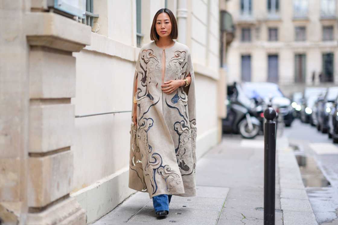 Aimee Song wears a long beige embroidered cape dress and blue denim pants