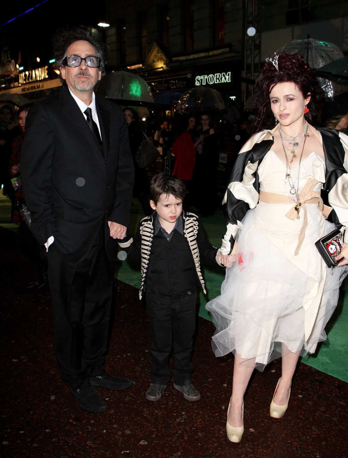 A man in a suit, a young boy in a striped jacket, and a woman in a white and black gown walk together on a rainy red carpet. A man in a suit, a young boy in a striped jacket, and a woman in a white and black gown walk together on a rainy red carpet.