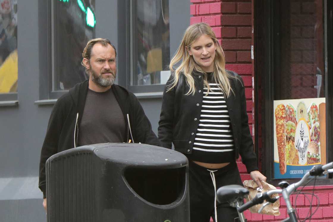 Jude Law in a dark hoodie walks beside a woman in a striped top past a bin and bicycle.
