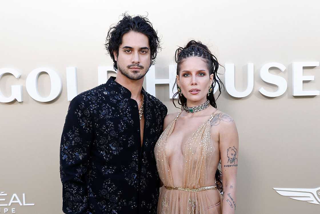 A couple poses at a step-and-repeat, the man in an embellished black shirt and the woman in a sheer gold dress with visible tattoos.