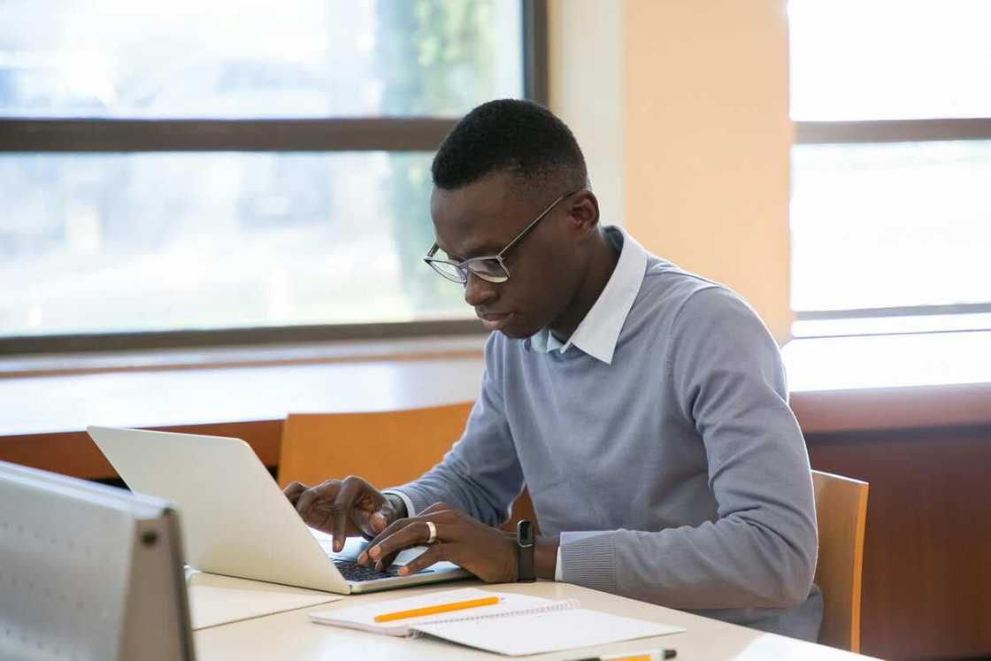 A young man is working on a laptop sitting near the window in the library.