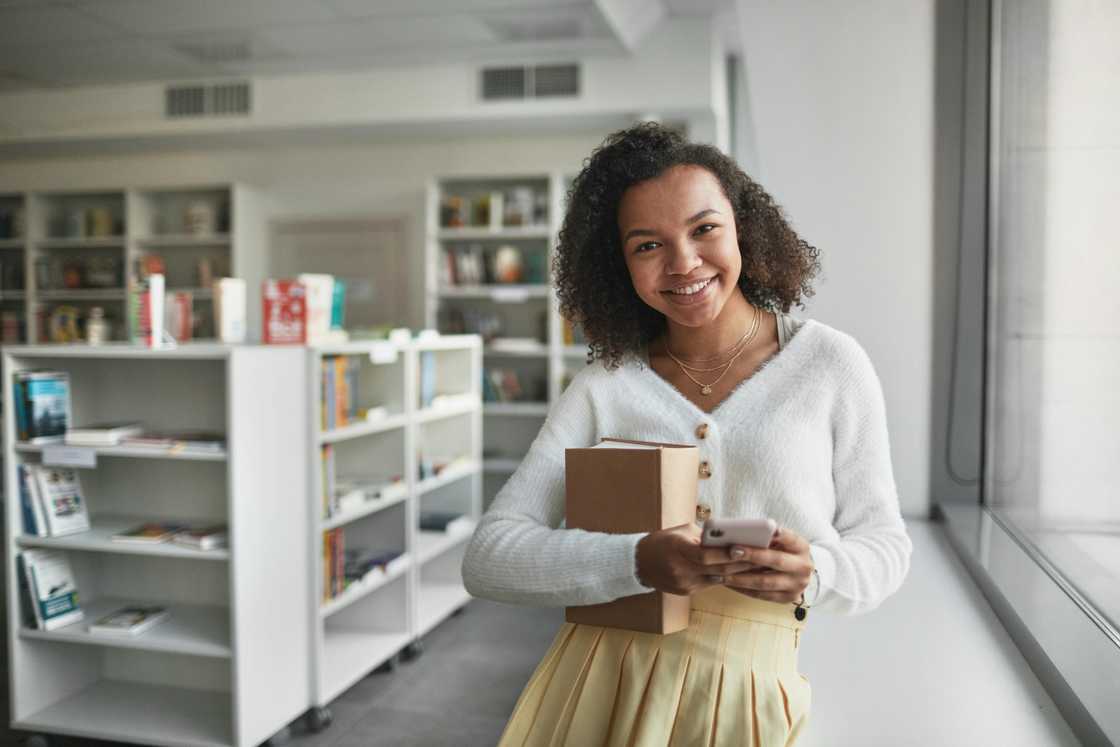 A smiling young woman in a white top and brown skirt is holding a book