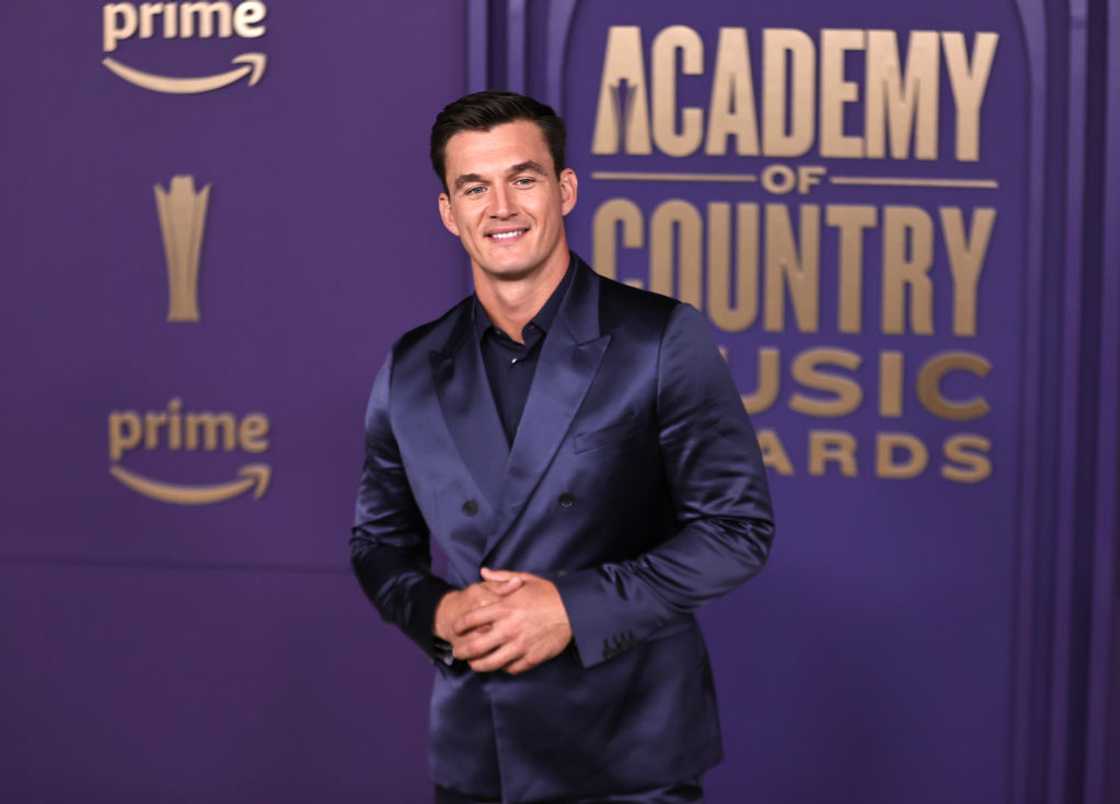 Tyler Cameron poses in a shiny navy suit in front of an Academy of Country Music Awards backdrop.